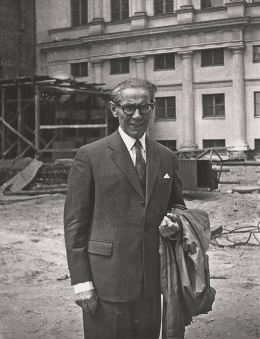 Na czarno białym zdjęciu Arnold Szyfman stojący przed budynkiem Teatru Wielkiego w Warszawie, z płaszcze zawieszonym na ręce.
